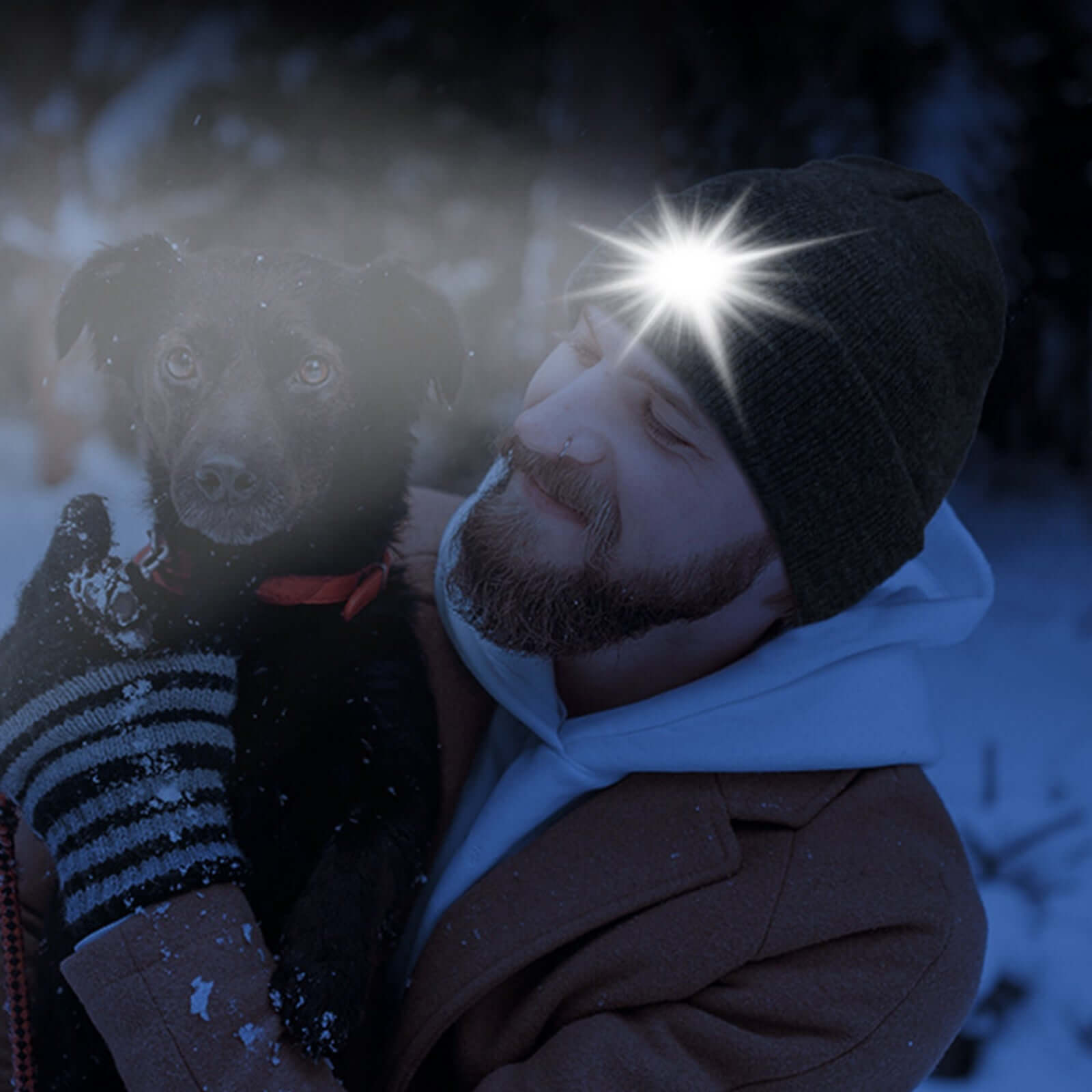 Man wearing knit beanie hat with built-in LED light holding a dog in winter forest